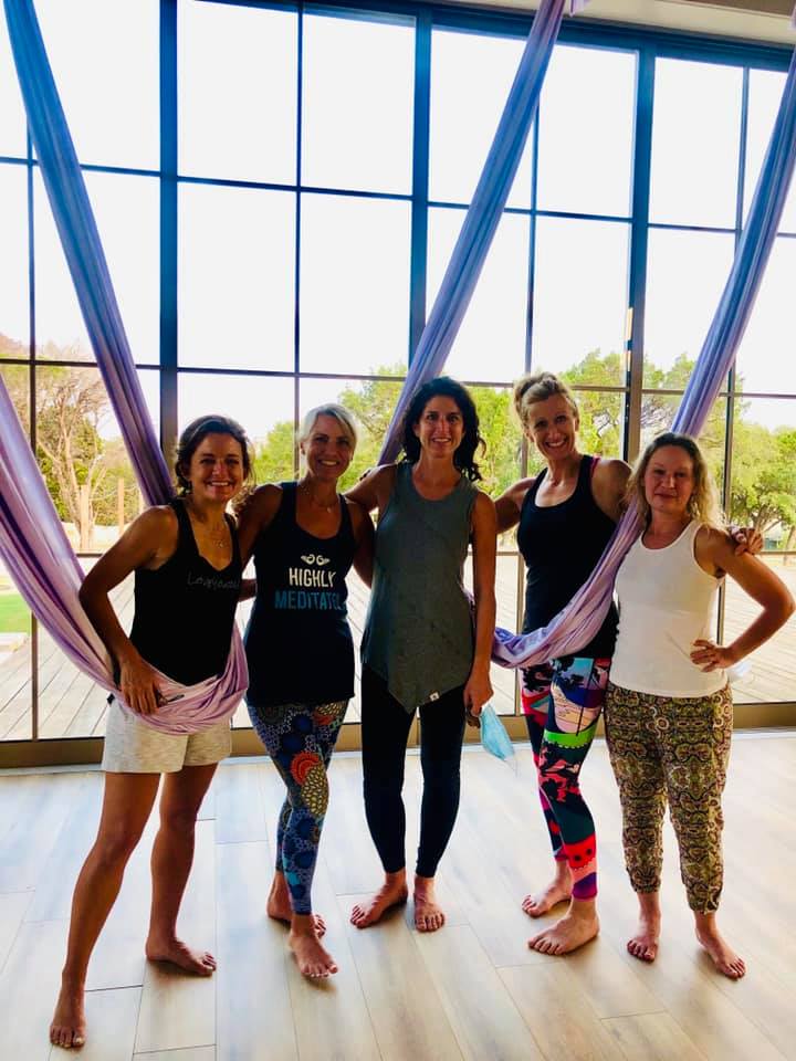 Five women in athletic wear posing inside a bright, spacious yoga studio.