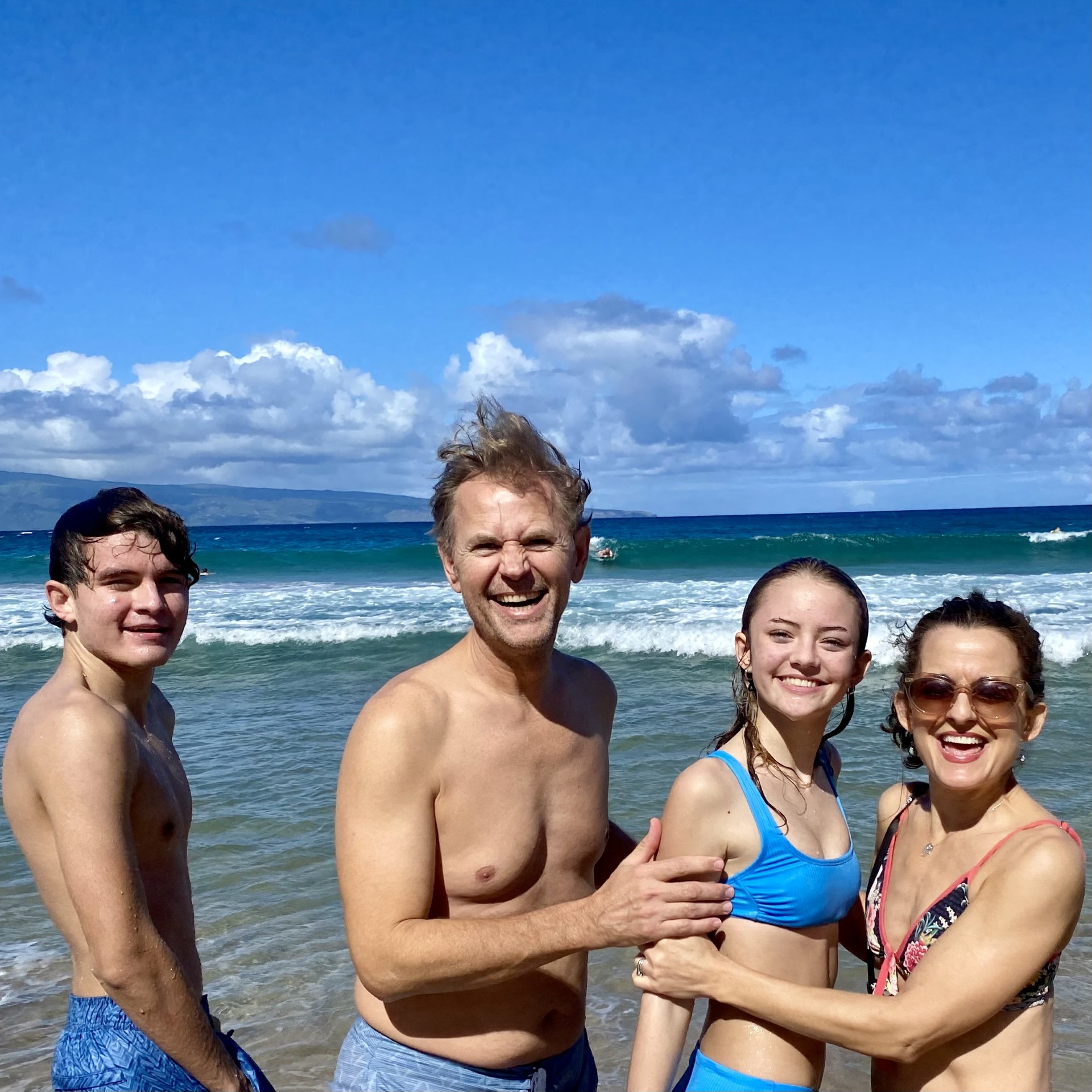 Four friends smiling and posing shirtless at a sunny beach with waves behind them.